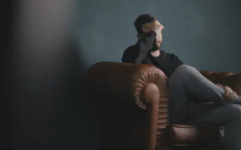Άνδρας σκυφτός σε δερμάτινη πολυθρόνα με το χέρι στο πρόσωπο, σε σκοτεινό δωμάτιο.