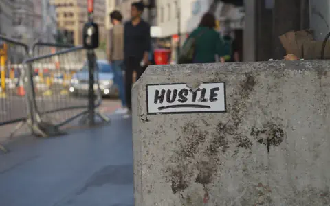 Πινακίδα με τη λέξη HUSTLE πάνω σε τσιμεντένιο εμπόδιο σε δρόμο πόλης.