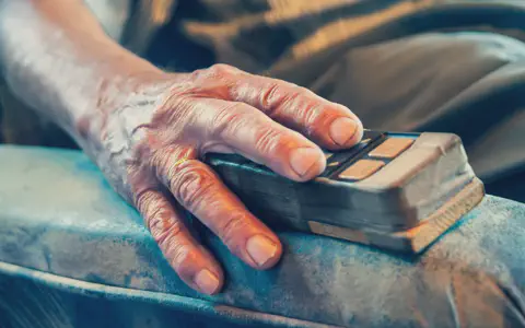 Χέρι ηλικιωμένου που κρατά μια παλιά συσκευή πάνω σε μπλε ύφασμα.
